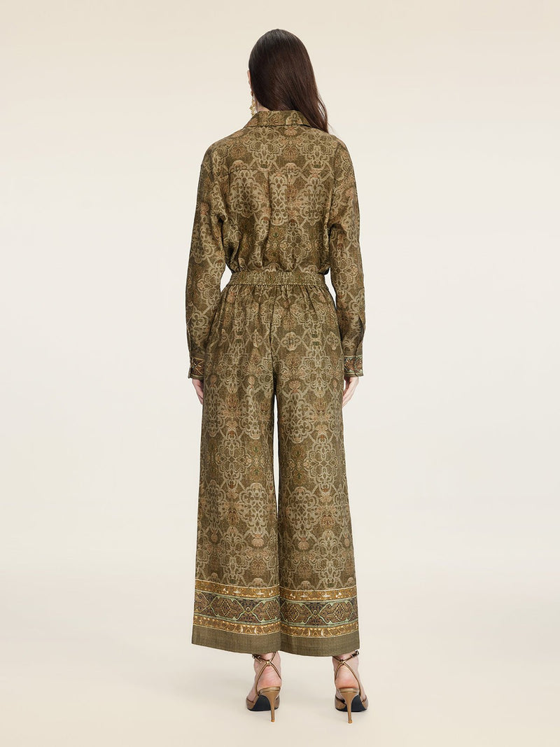 A person stands facing away, wearing Social Queen's Printed Linen Straight-Leg Pants in olive green with gold detailing, paired with a matching patterned long-sleeve top and open-toe heels.