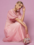 A woman in a Puff Sleeve Textured Lace Dress by Social Queen sits against a pink background, holding a small floral handbag and wearing statement earrings.