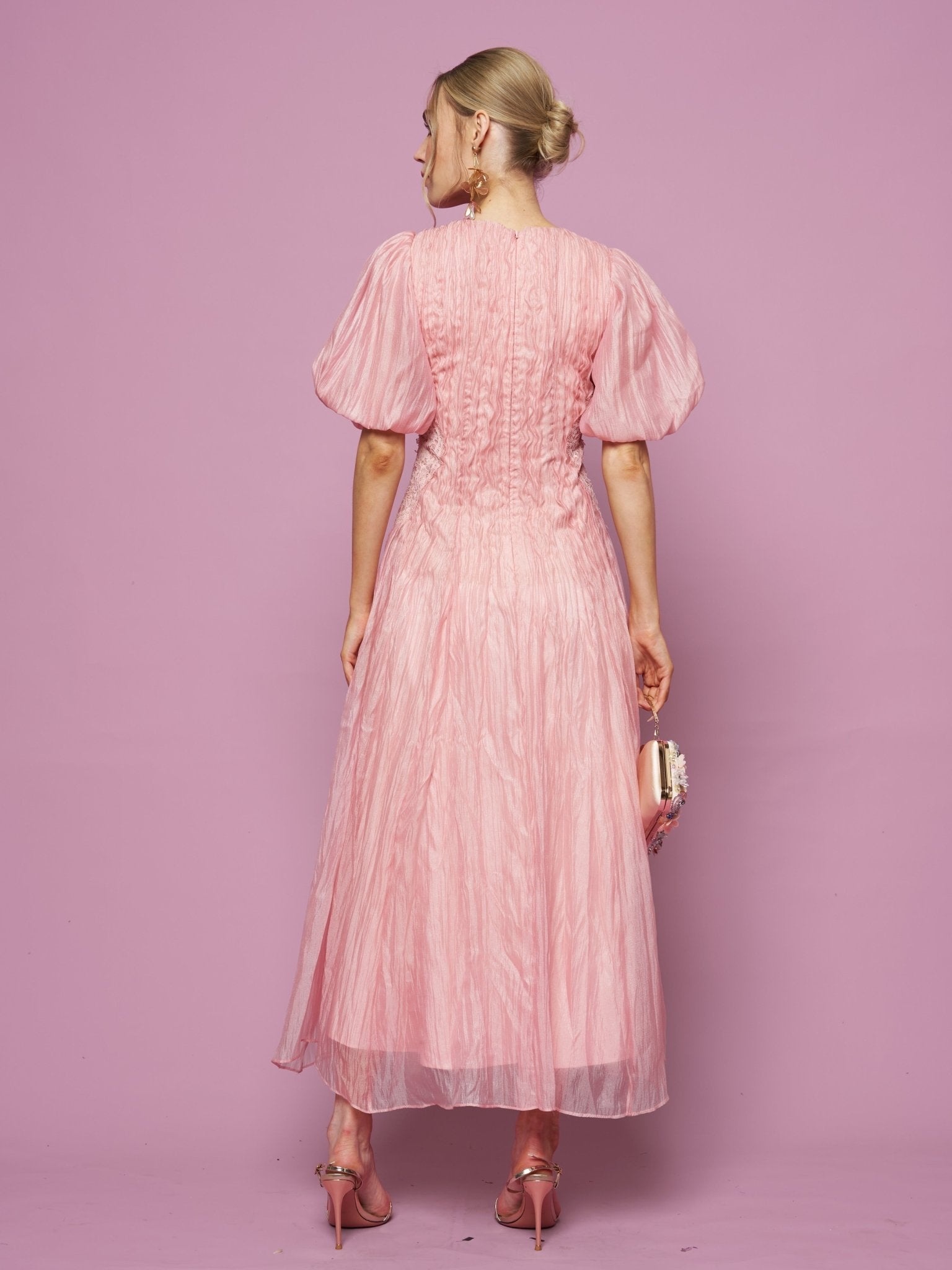 A woman faces away from the camera in a Social Queen Puff Sleeve Textured Lace Dress, paired with pink heels and a small handbag, set against a pink backdrop.