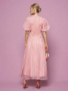 A woman faces away from the camera in a Social Queen Puff Sleeve Textured Lace Dress, paired with pink heels and a small handbag, set against a pink backdrop.