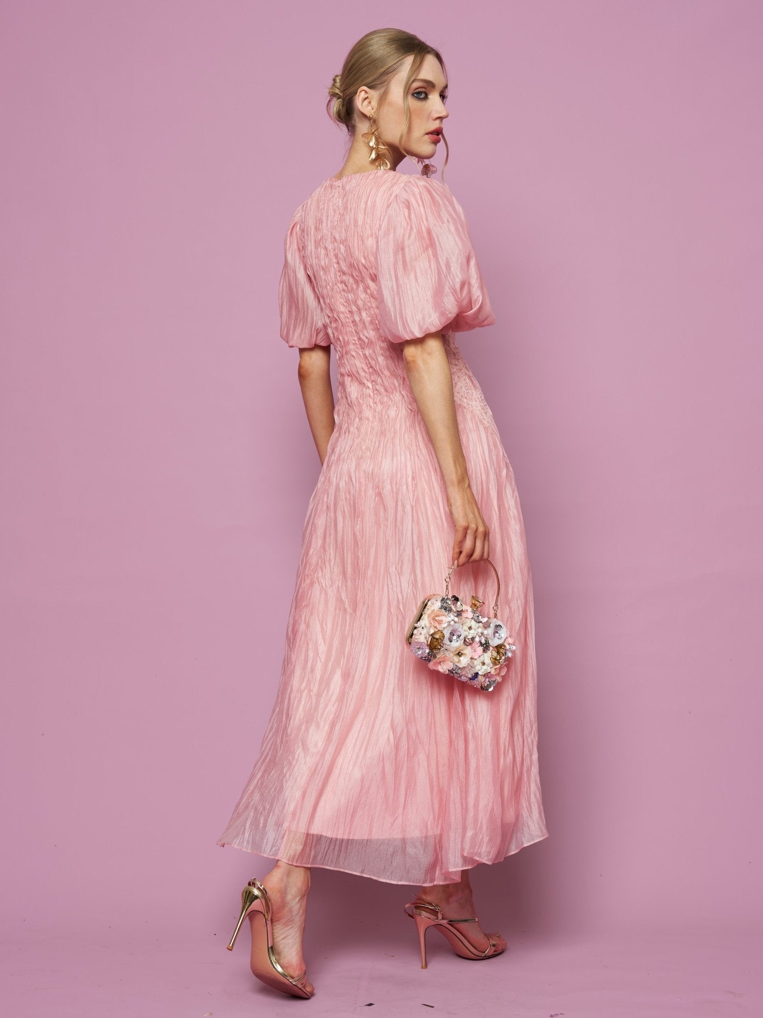 A woman stands against a pink background, wearing the Social Queen Puff Sleeve Textured Lace Dress with metallic heels, holding a floral handbag, and looking over her shoulder.