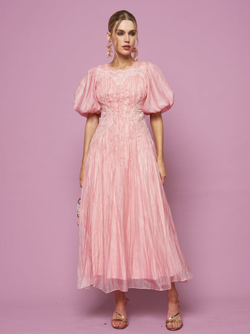 A woman stands against a pink background, wearing the Social Queen Puff Sleeve Textured Lace Dress in light pink, styled with earrings and strappy sandals.