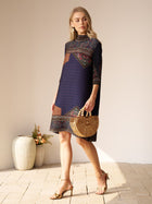 A woman wearing the Social Queen Retro Ethnic Print Pleated Dress and sandals holds a wooden handbag while standing indoors near vases with flowers.