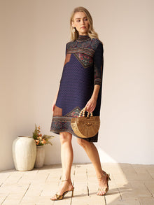A woman wearing the Social Queen Retro Ethnic Print Pleated Dress and sandals holds a wooden handbag while standing indoors near vases with flowers.
