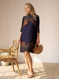 A woman indoors stands on a tiled floor, holding a wooden handbag beside a wicker chair, wearing the Social Queen Retro Ethnic Print Pleated Dress as sunlight streams in.