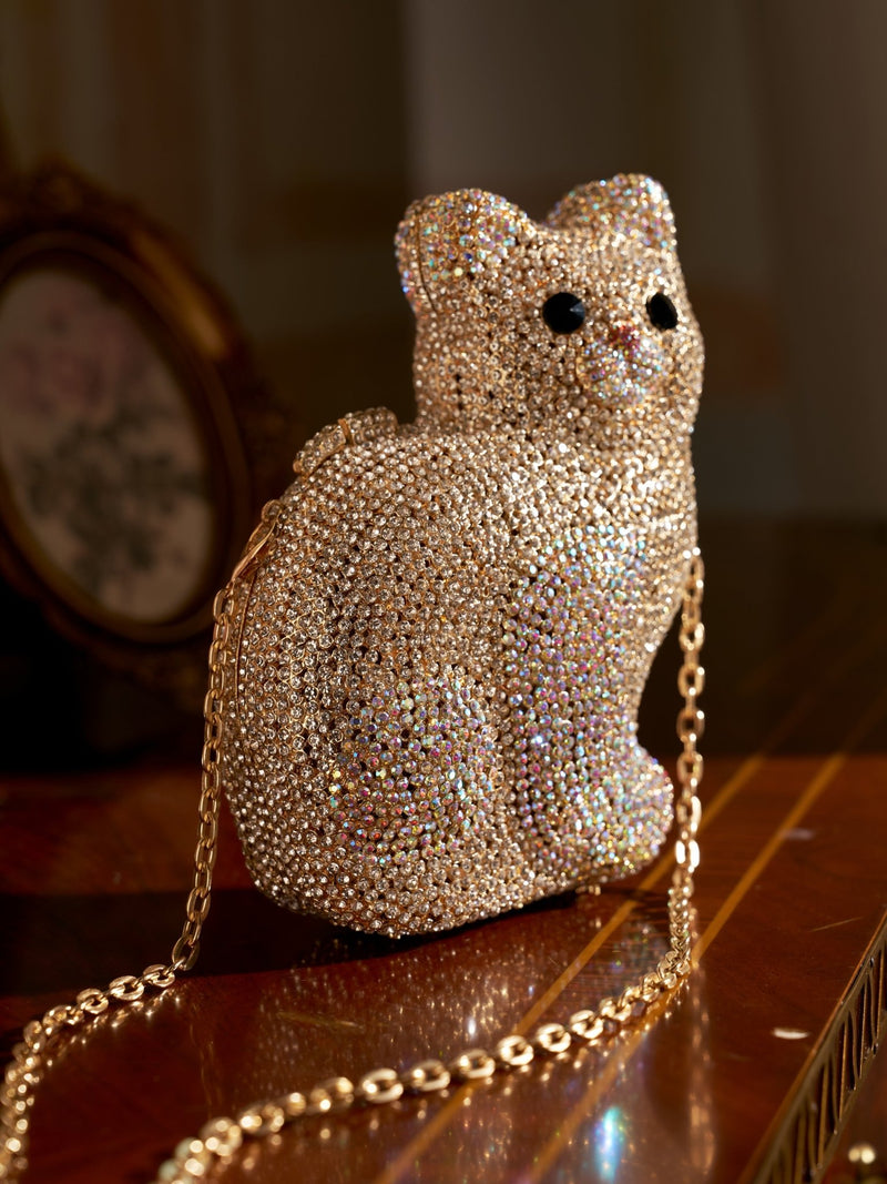 The Social Queen Rhinestone Cat Hardcase Clutch, featuring a sparkling design and gold chain strap, rests on a wooden surface—ideal as an evening bag or crossbody purse for special occasions.