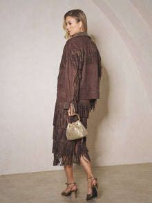 Woman poses indoors in the Social Queen Rhinestone Fringe Suede Midi Skirt, paired with metallic heels and a small gold handbag, looking over her shoulder.