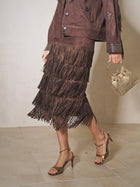 A person wearing the Social Queen Rhinestone Fringe Suede Midi Skirt, paired with a matching studded jacket, gold heeled sandals, and a small gold handbag.