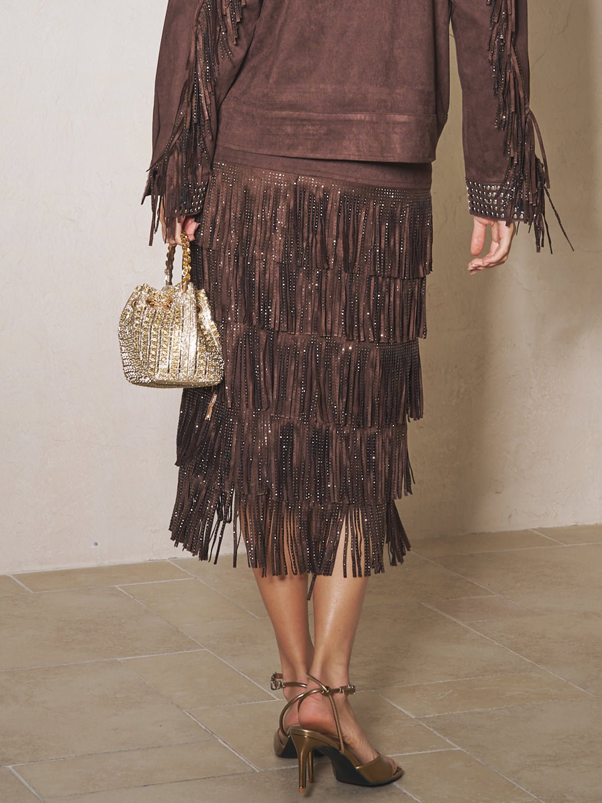 A person wears the Social Queen Rhinestone Fringe Suede Midi Skirt with a matching jacket, holds a small gold handbag, and pairs the look with brown heeled sandals.