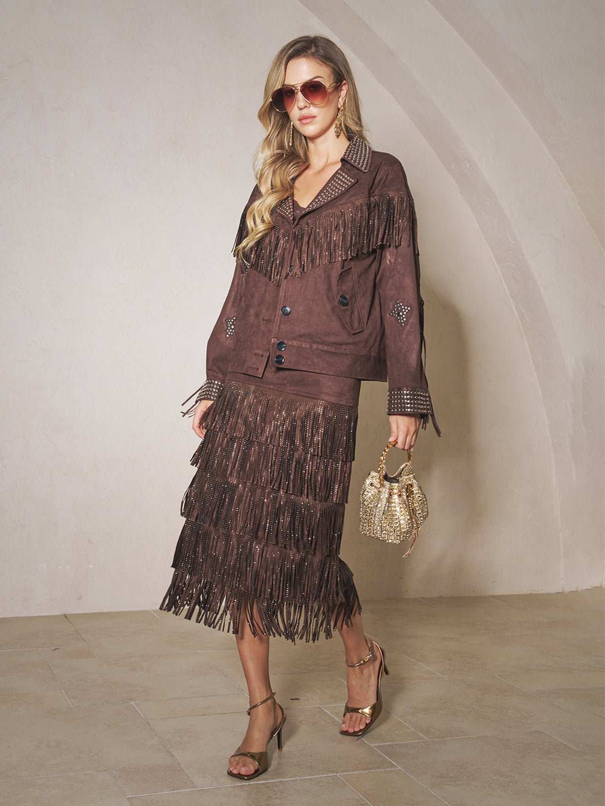 A woman wearing the Social Queen Rhinestone Fringe Suede Midi Skirt, a matching jacket, sunglasses, heeled sandals, and holding a small gold handbag poses indoors against a neutral wall.