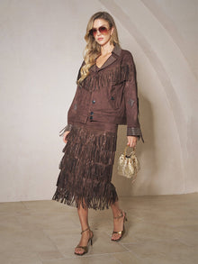 A woman wearing the Social Queen Rhinestone Fringe Suede Midi Skirt, a matching jacket, sunglasses, heeled sandals, and holding a small gold handbag poses indoors against a neutral wall.