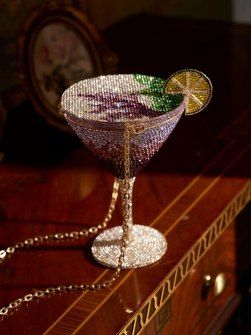 The Social Queen Rhinestone Martini Glass Hardcase Clutch, adorned with a jeweled lime and gold chain strap, rests on a wooden table—your perfect crossbody party purse for any glamorous night.