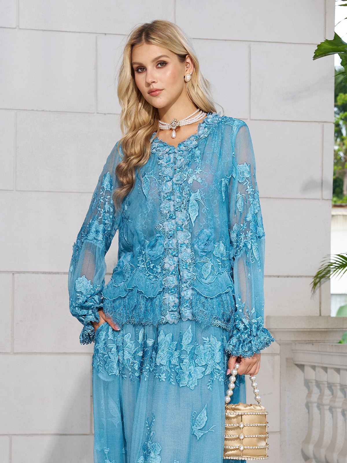 A woman in the Social Queen Rose Appliqué 100% Mulberry Silk Blouse with romantic puff sleeves and a matching skirt stands against a white stone wall, holding a beige beaded handbag and wearing a choker necklace.