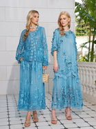 Two women stand side by side in light blue Rose Appliqué 100% Mulberry Silk Blouses by Social Queen, styled with romantic puff sleeves and sandals, posed on a tiled patio with a white stone wall and greenery behind them.