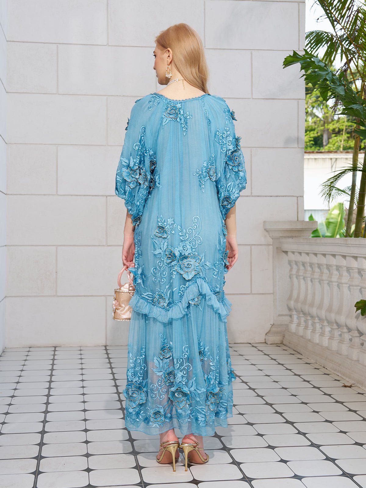 A woman with blonde hair stands facing away, wearing the Social Queen Rose Appliqué 100% Mulberry Silk Dress in light blue and holding a small beige handbag, on a tiled floor near a white wall and railing.