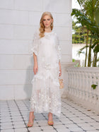A woman stands on a tiled patio in front of a white wall, wearing the Social Queen Rose Appliqué 100% Mulberry Silk Dress with sheer floral details, gold heels, and holding a small handbag.