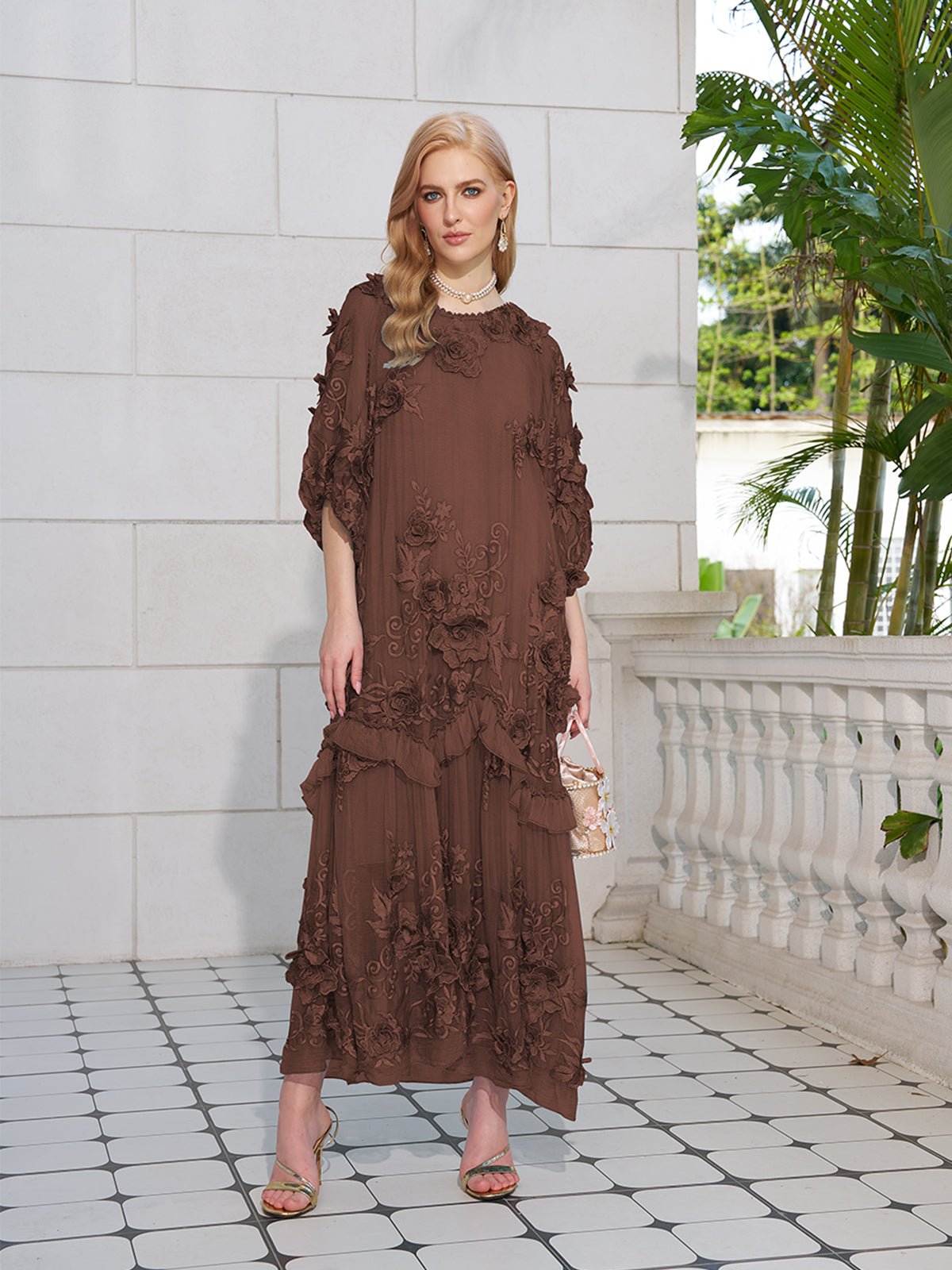 A woman wears the Social Queen Rose Appliqué 100% Mulberry Silk Dress, a long brown A-line with floral accents, as she stands on a tiled patio near a white stone railing and lush green plants.