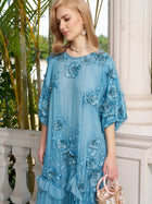 A woman wearing the Social Queen Rose Appliqué 100% Mulberry Silk Dress, delicately floral-embroidered in light blue, stands by a white column with greenery, holding a small handbag and looking down.