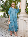 A woman in Social Queen's loose, blue Rose Appliqué 100% Mulberry Silk Dress stands on a tiled veranda, holding a small handbag, with greenery and white pillars behind her.
