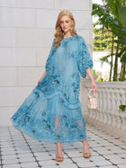 A woman in a light blue Rose Appliqué 100% Mulberry Silk Dress by Social Queen stands on a tiled patio, holding a small handbag, with plants and a white stone wall behind her.