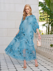 A woman in a light blue Rose Appliqué 100% Mulberry Silk Dress by Social Queen stands on a tiled patio, holding a small handbag, with plants and a white stone wall behind her.