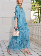 A woman in the Social Queen Rose Appliqué 100% Mulberry Silk Dress holds a light handbag and wears gold heels, standing on tiled flooring beside a white balustrade.
