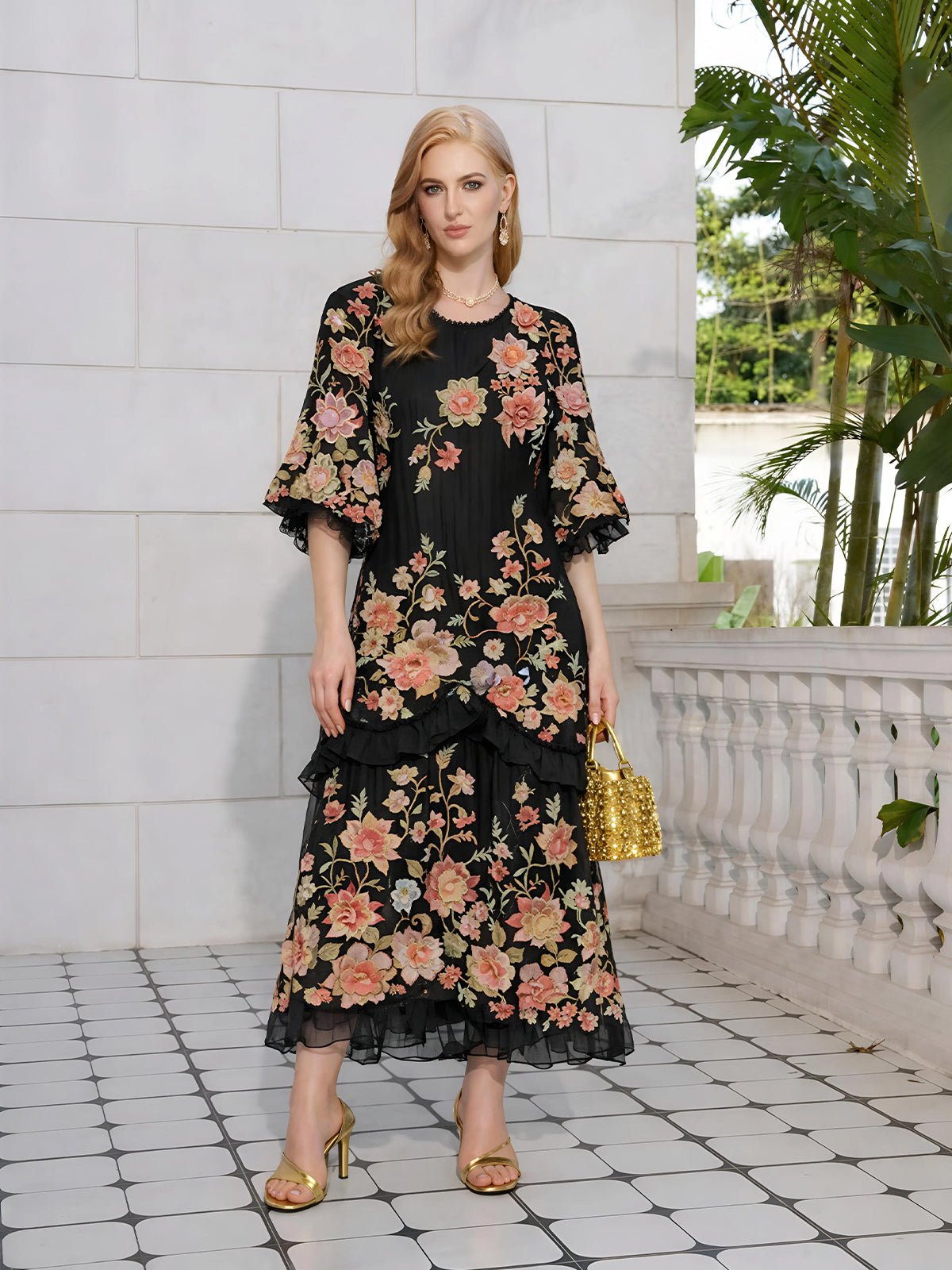 A woman stands on a tiled patio wearing the Social Queen Rose Appliqué 100% Mulberry Silk Dress with colorful floral embroidery, gold sandals, and a gold handbag. Green plants and a white stone railing are in the background.