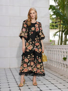 A woman stands on a tiled patio wearing the Social Queen Rose Appliqué 100% Mulberry Silk Dress with colorful floral embroidery, gold sandals, and a gold handbag. Green plants and a white stone railing are in the background.