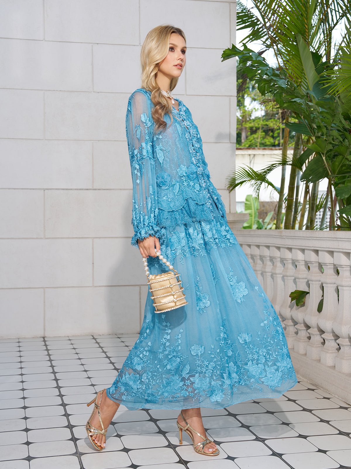 A woman in the Social Queen Rose Appliqué 100% Mulberry Silk Skirt and strappy heels holds a small woven handbag, standing on a tiled patio surrounded by plants.