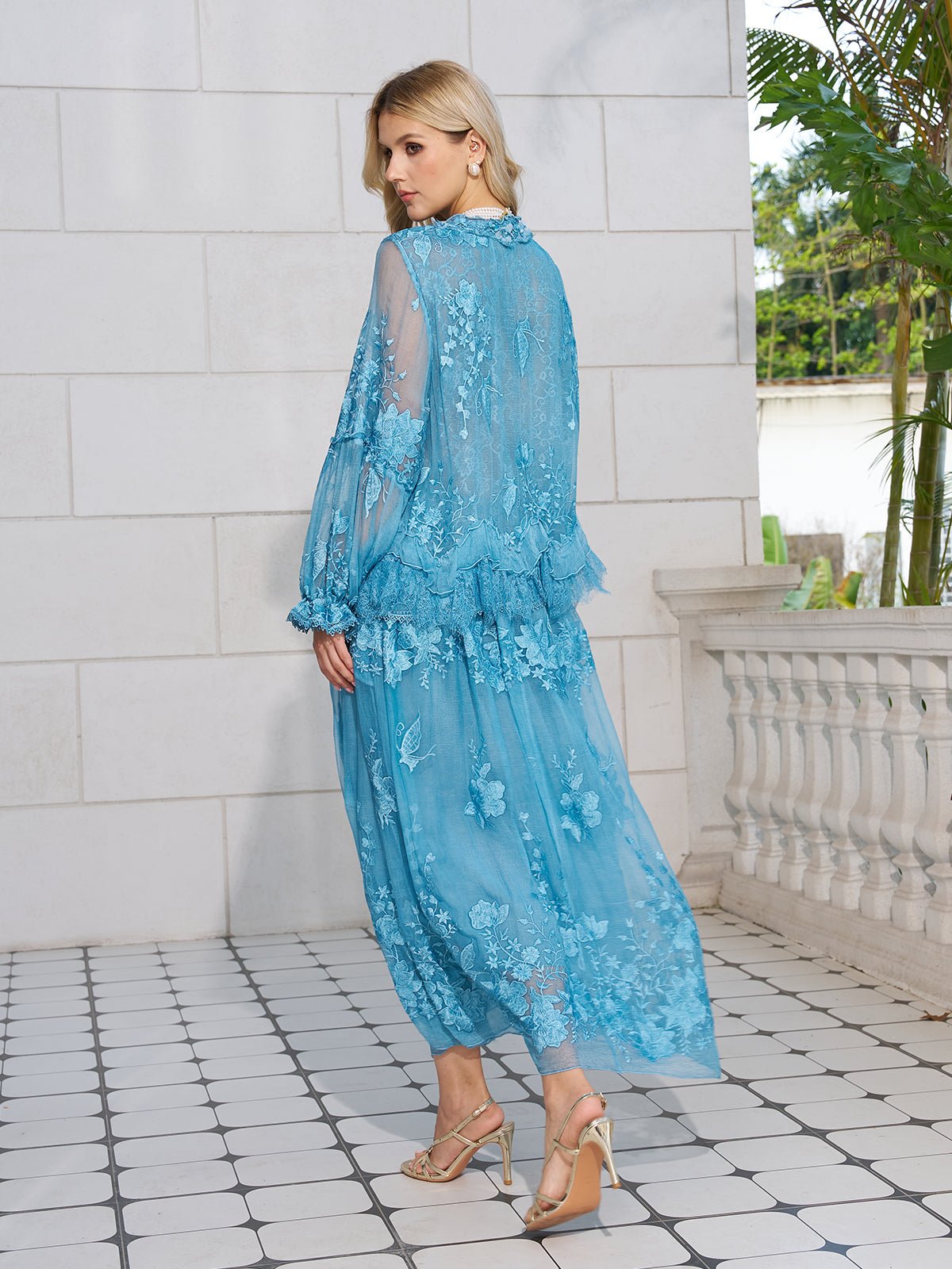 A woman stands on a tiled patio, facing away, wearing the Social Queen Rose Appliqué 100% Mulberry Silk Skirt with floral details and heeled sandals.