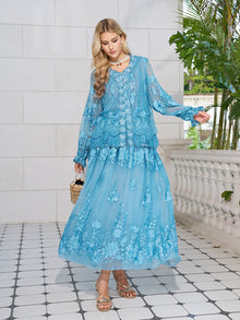 A woman stands on a tiled patio wearing the Social Queen Rose Appliqué 100% Mulberry Silk Skirt in light blue, paired with sandals, a necklace, and a small woven handbag. Green plants decorate the background.