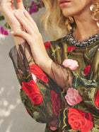 A woman wears the Social Queen Rose Embroidered Beaded A-Line Dress with sheer sleeves, large gold earrings, and pink flowers in the background—a chic choice for a stylish wedding guest.