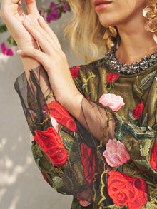A woman wears the Social Queen Rose Embroidered Beaded A-Line Dress with sheer sleeves, large gold earrings, and pink flowers in the background—a chic choice for a stylish wedding guest.