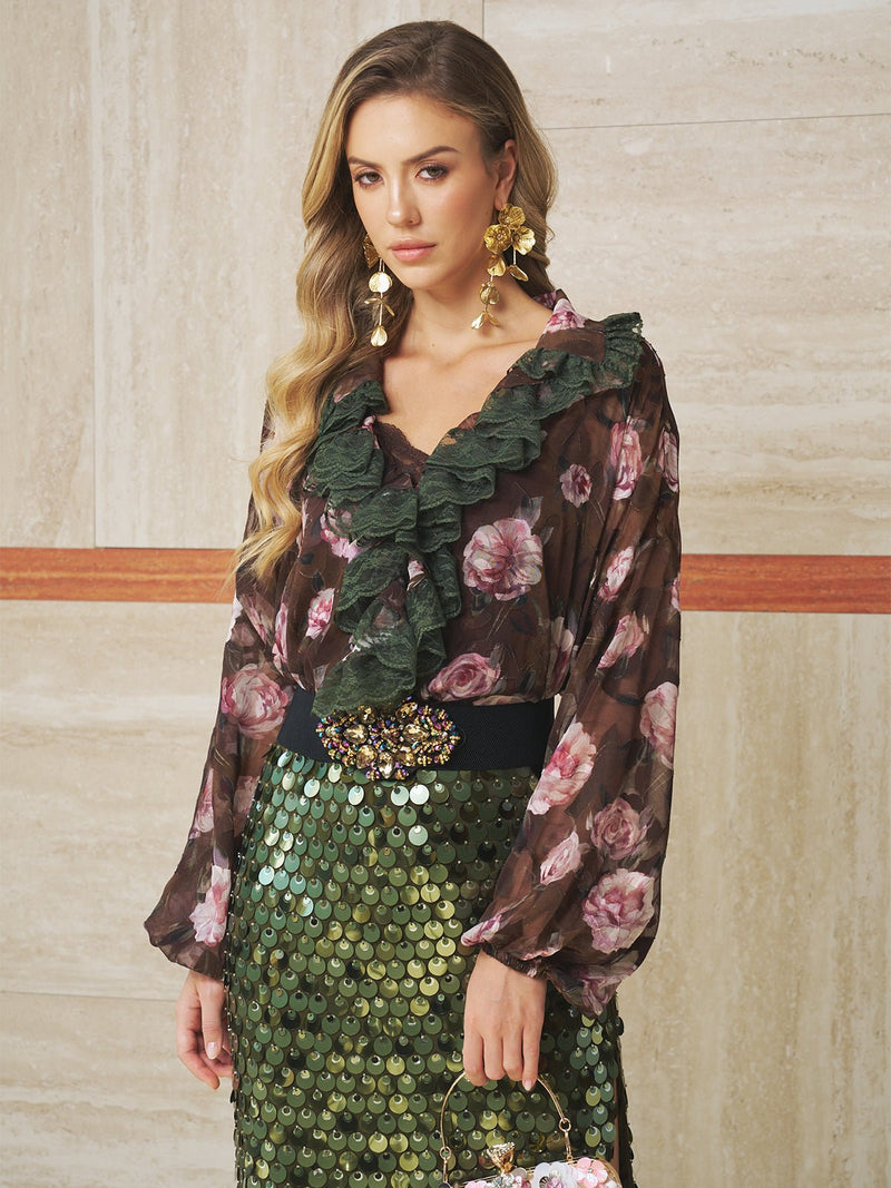 A woman wearing the Social Queen Rose Jacquard Lace Ruffle Blouse, a green sequin skirt, and large gold earrings stands against a beige marble wall.