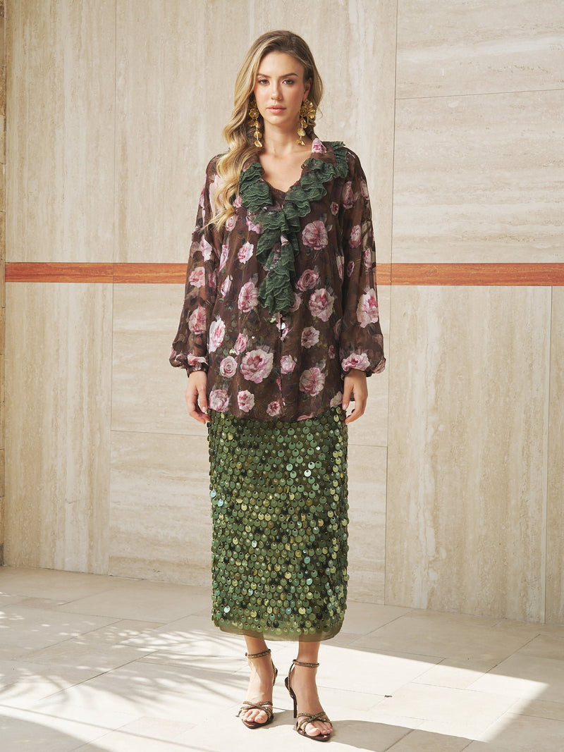 A woman stands indoors wearing the Social Queen Rose Jacquard Lace Ruffle Blouse, paired with a green textured skirt, strappy sandals, and statement earrings.