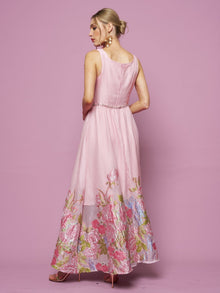 A woman with blonde hair in a bun models the Social Queen Rose Jacquard Sleeveless Gown, featuring floral embroidery at the hem, as she stands against a pink background facing away from the camera.