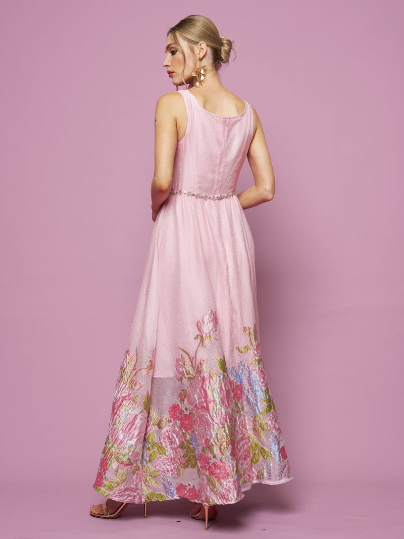 A woman with blonde hair in a bun models the Social Queen Rose Jacquard Sleeveless Gown, featuring floral embroidery at the hem, as she stands against a pink background facing away from the camera.
