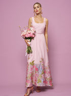 A woman wearing the Social Queen Rose Jacquard Sleeveless Gown holds a bouquet of pink flowers, standing against a solid pink background.