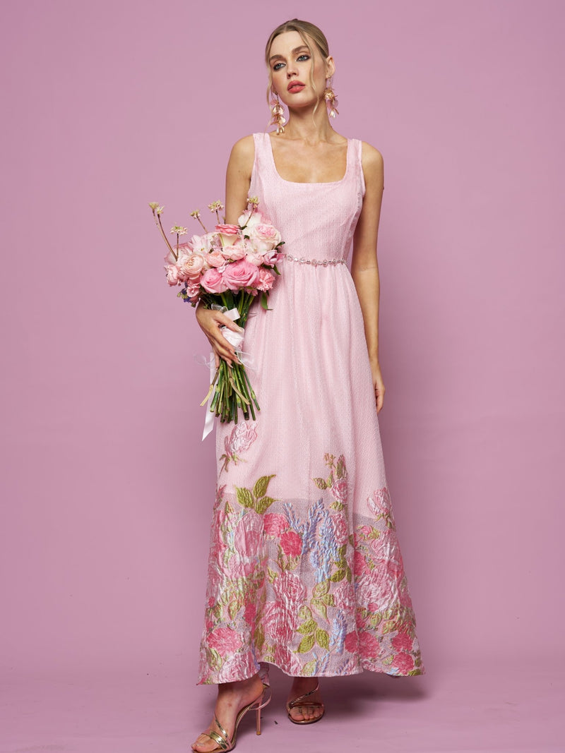 A woman wearing the Social Queen Rose Jacquard Sleeveless Gown holds a bouquet of pink flowers, standing against a solid pink background.