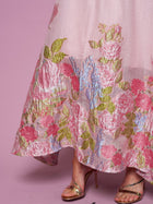 A person in the Rose Jacquard Sleeveless Gown by Social Queen, featuring a metallic sheen and crystal trim, paired with gold strappy high heel sandals, stands against a pink background.