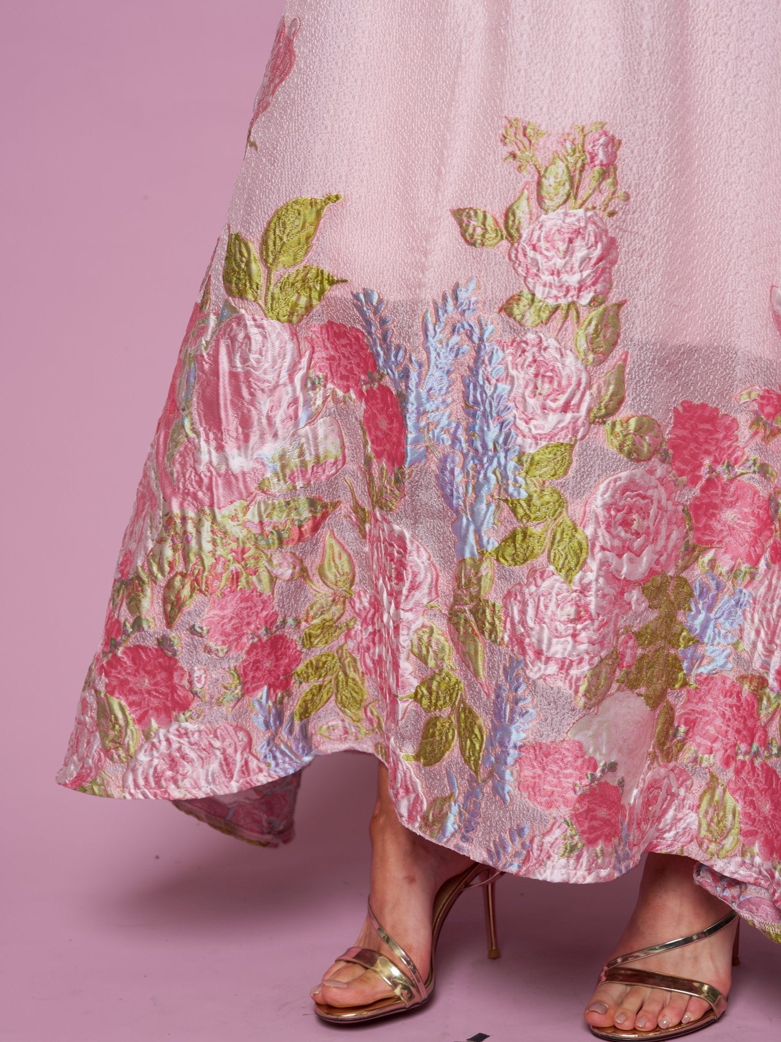 A person in the Rose Jacquard Sleeveless Gown by Social Queen, featuring a metallic sheen and crystal trim, paired with gold strappy high heel sandals, stands against a pink background.