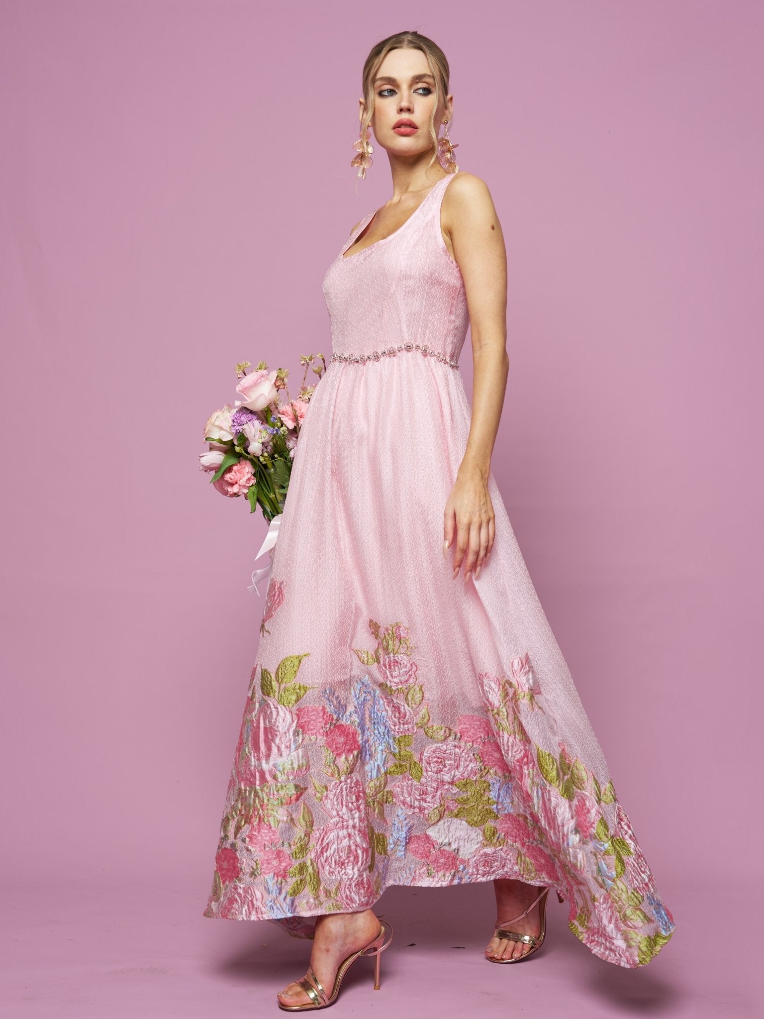 A woman poses elegantly against a pink backdrop in the Social Queen Rose Jacquard Sleeveless Gown with crystal-trim detail, holding a bouquet of flowers.