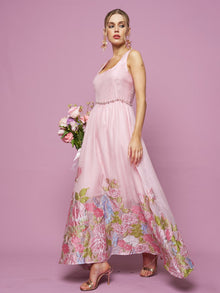 A woman poses elegantly against a pink backdrop in the Social Queen Rose Jacquard Sleeveless Gown with crystal-trim detail, holding a bouquet of flowers.