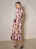 A woman models the Social Queen Rose Print Knit Dress, featuring pink floral patterns and long sleeves, paired with a gold handbag and high-heeled sandals, standing sideways against a plain, light background.