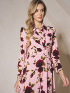 A woman in the Social Queen Rose Print Knit Dress, featuring red and black roses and long sleeves, stands against a beige wall, gazing slightly to the side.