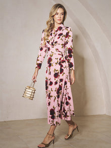 A woman in the Rose Print Knit Dress by Social Queen holds a small handbag and wears gold high-heeled sandals, standing indoors against a light wall.