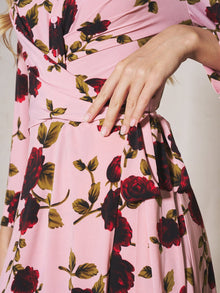 A person wears the Social Queen Rose Print Knit Dress, a pink French-inspired piece with red and green floral accents, resting their hand on their waist.