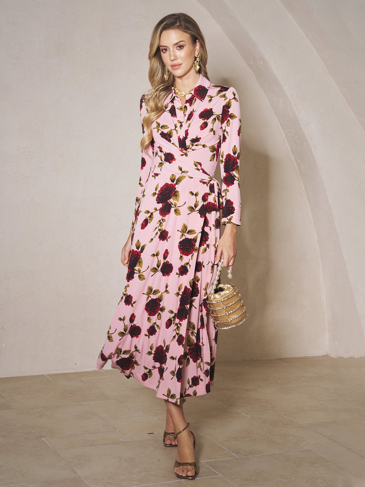 A woman stands indoors wearing the Social Queen Rose Print Knit Dress, featuring long sleeves and a red and black floral pattern, paired with heeled sandals and a small handbag.