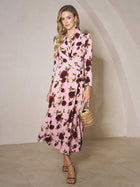 A woman stands indoors wearing the Social Queen Rose Print Knit Dress, featuring long sleeves and a red and black floral pattern, paired with heeled sandals and a small handbag.