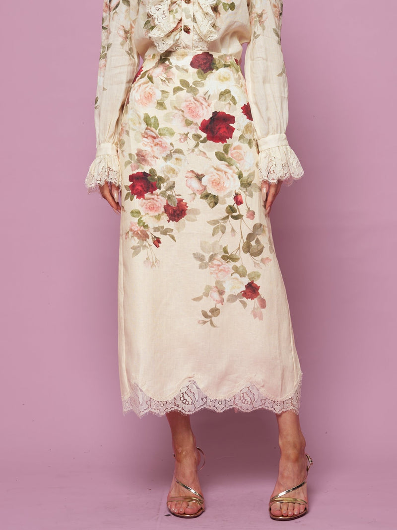 A person stands against a pink background wearing the Social Queen Rose Print Lace Hem Linen Skirt paired with a matching blouse and silver strappy heels.